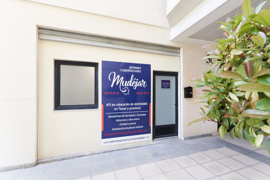 Puerta de la oficina de construcciones mudejar con cartel grande en colores azul, blanco y rojo, una ventana y una puerta acristalada opaca