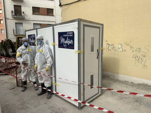 Dos trabajadores protegidos con traje blanco profesional y mascarilla delante de caseta de construcción