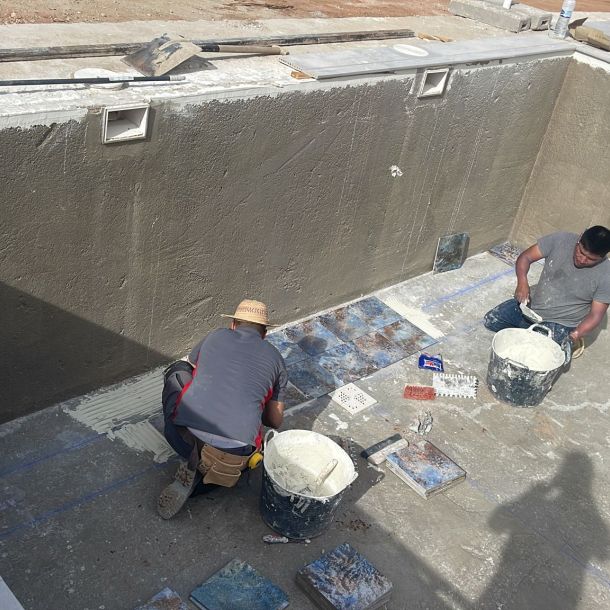 Dos profesionales dentro de una piscina de construcción poniendo baldosas 