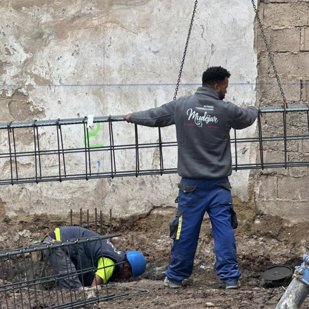 Obrero con sudadera de construcciones mudéjar moviendo con las manos una estructura metálica con la ayuda de una grúa 