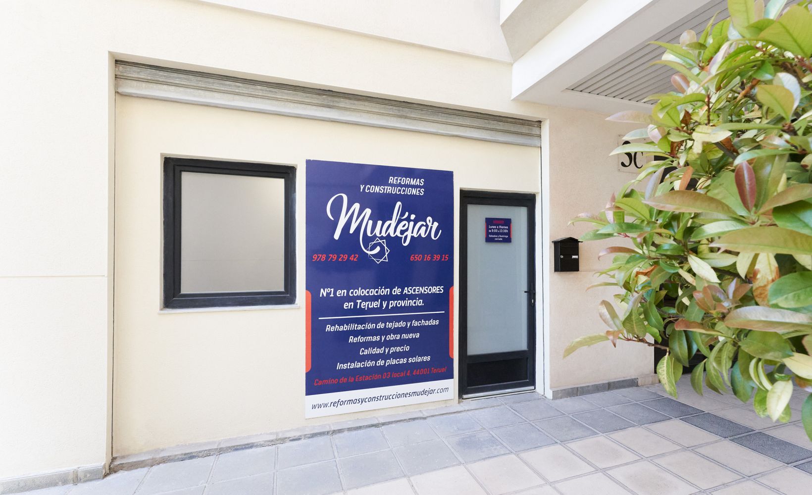 Imagen de la fachada exterior de la oficina de construcciones mudéjar con cartel de color azul, rojo y blanco y ventana y puerta de cristal opacas 
